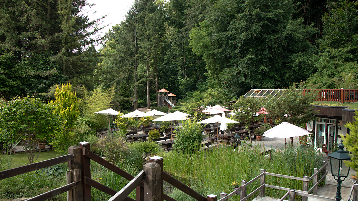 Pohled na restauraci Boltenmühle u 53.0372067, 12.8039617 v Ruppinském Švýcarsku s terasou, slunečníky a lesní krajinou.